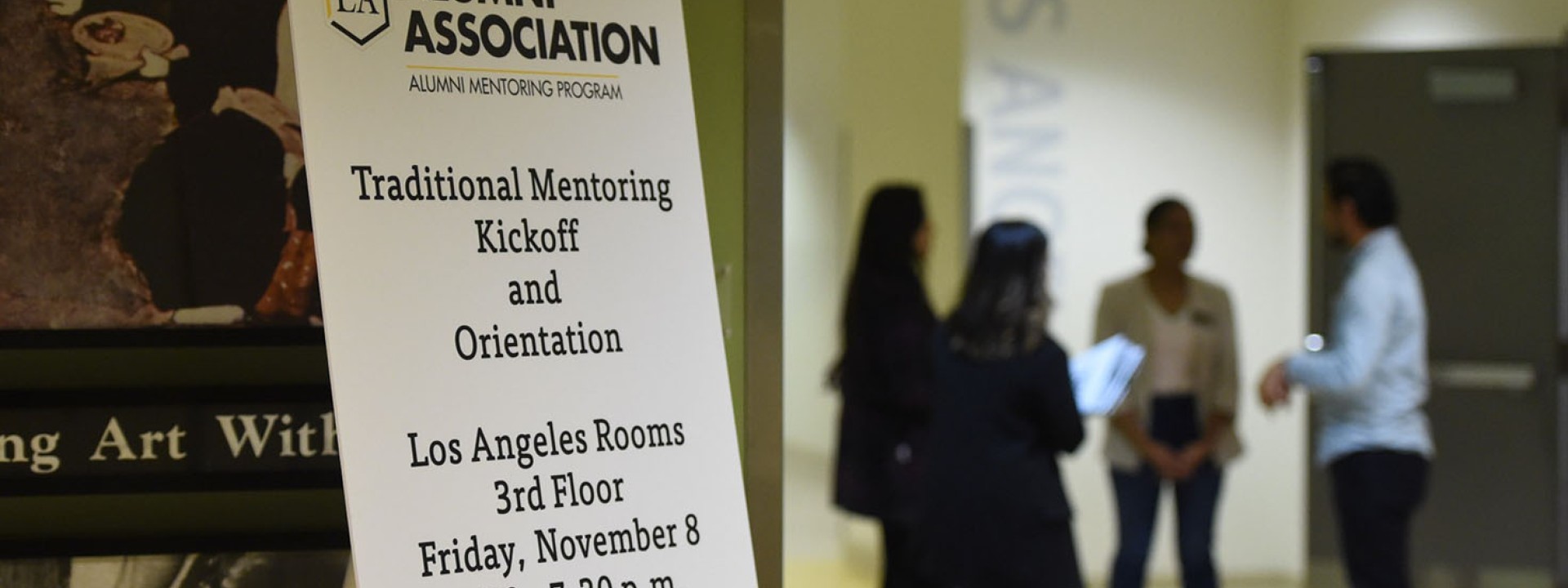 Sign for mentoring orientation with a group of people chatting in the background