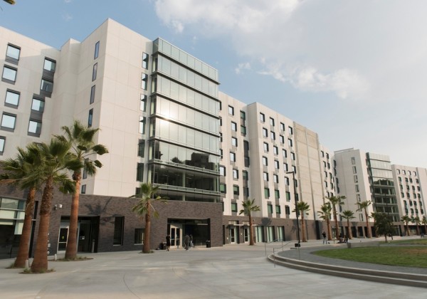 Modern student housing building at Cal State LA, with tall palm trees lining a wide concrete pathway and large glass windows reflecting the afternoon sun under a partly cloudy sky.