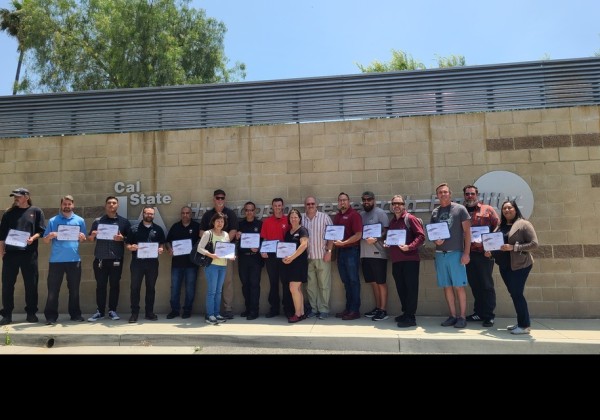Group of workshop participants holding certificates outside the Cal State LA Hydrogen Research Facility, standing in a row against a tan brick wall under a clear blue sky.