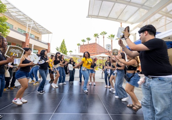 Students dance and cheer in a circle during Campus Day at Cal State LA, holding signs and waving flags in black and gold.