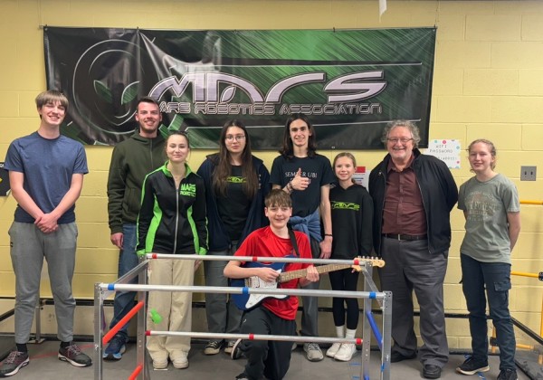 John Kennedy poses with students from the Mars Robotics Association in front of a banner and robot prototype.